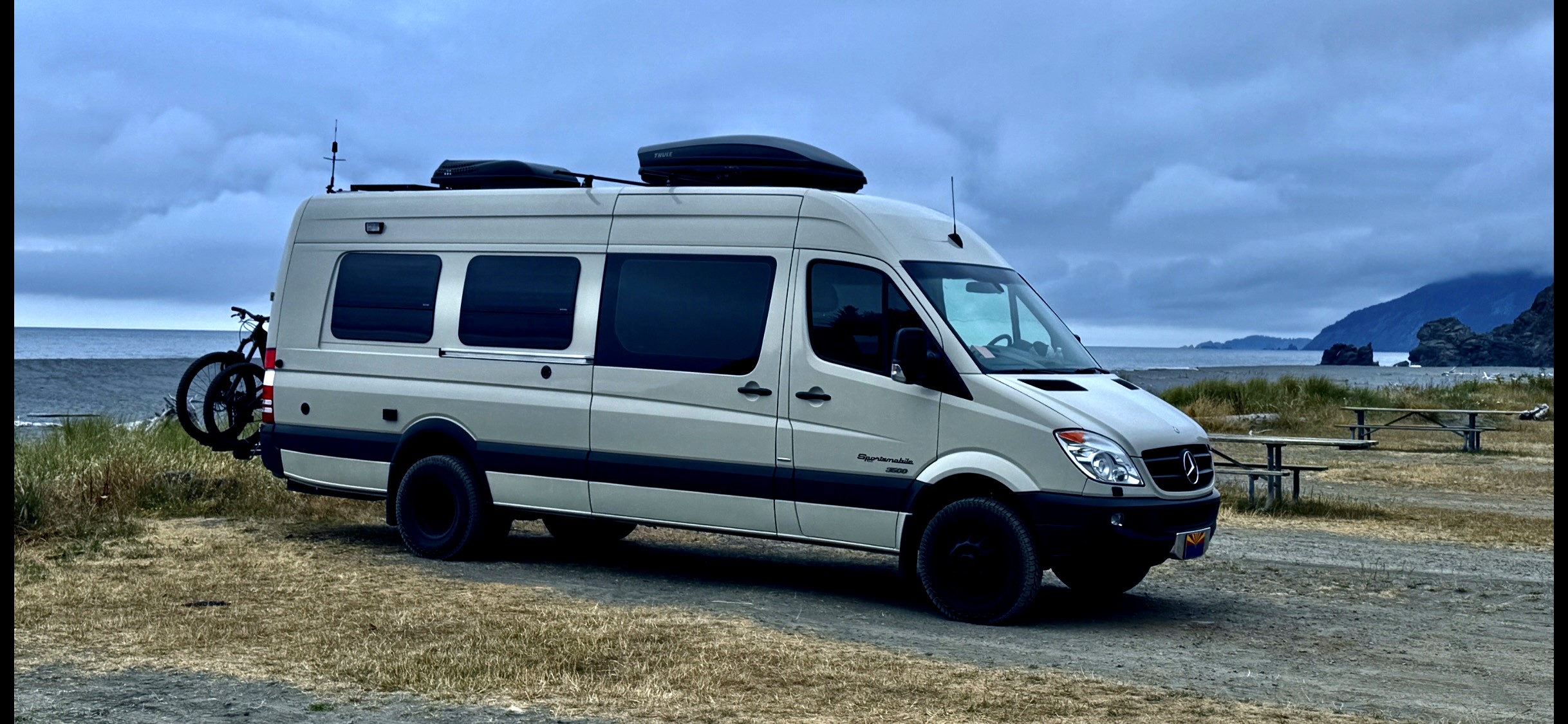A Sportsmobile Sprinter Class B camper van at a coastal campsite with bicycles on a rear rack